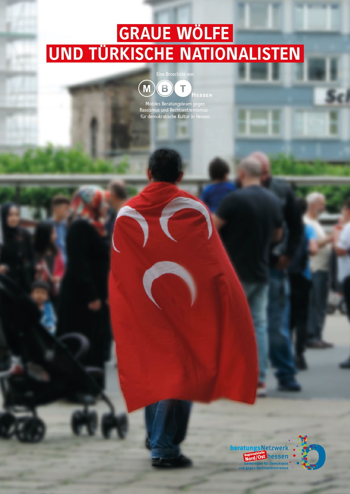 Auf dem Foto sieht man im Vordergrund eine Person auf einer Straße der eine mutmaßlich rote Fahne mit drei weißen Halbmonden trägt. Verschwommen im Hintergrund sieht man eine Menschenmenge die ebenfalls auf der Straße, vor Häusern steht. Auf dem Bild steht mit weißen Buchstaben auf roten Hintergrund geschrieben Graue Wölfe und türkische Nationalisten