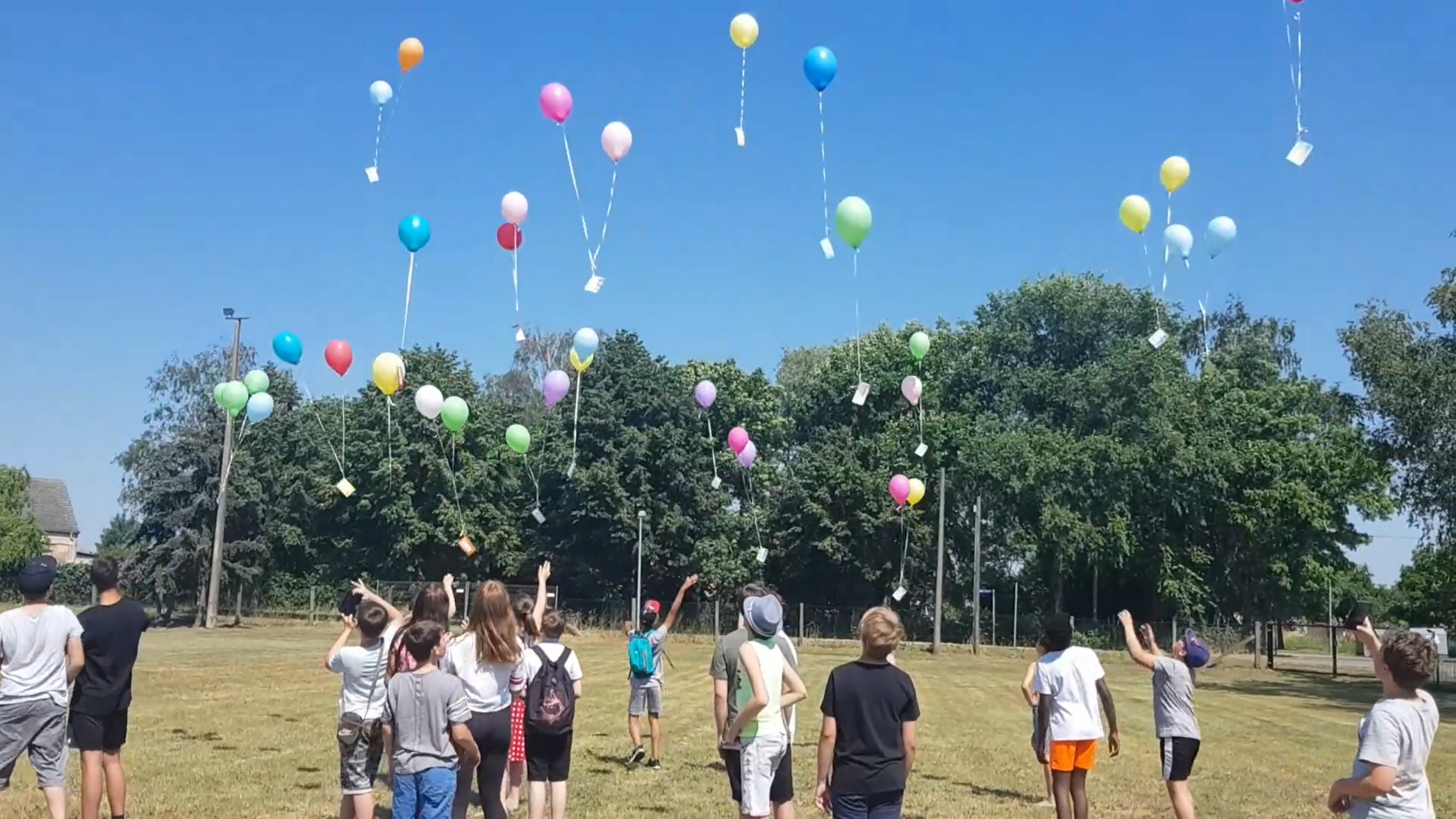 Auf dem Bild sieht man eine Gruppe von Kindern die auf einem Feld stehen und Luftballons mit Zetteln daran in den Himmel steigen lassen. Das Bild gehört zum Beitrag v