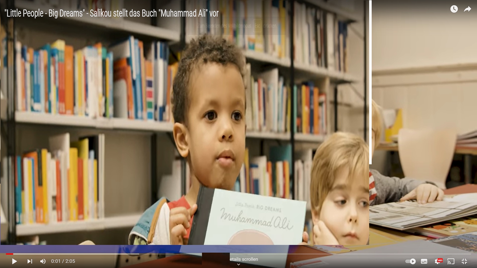 Ein schwarzer Junge steht vor einem Bücherregal und zeigt das Kinderbuch Muhammad Ali