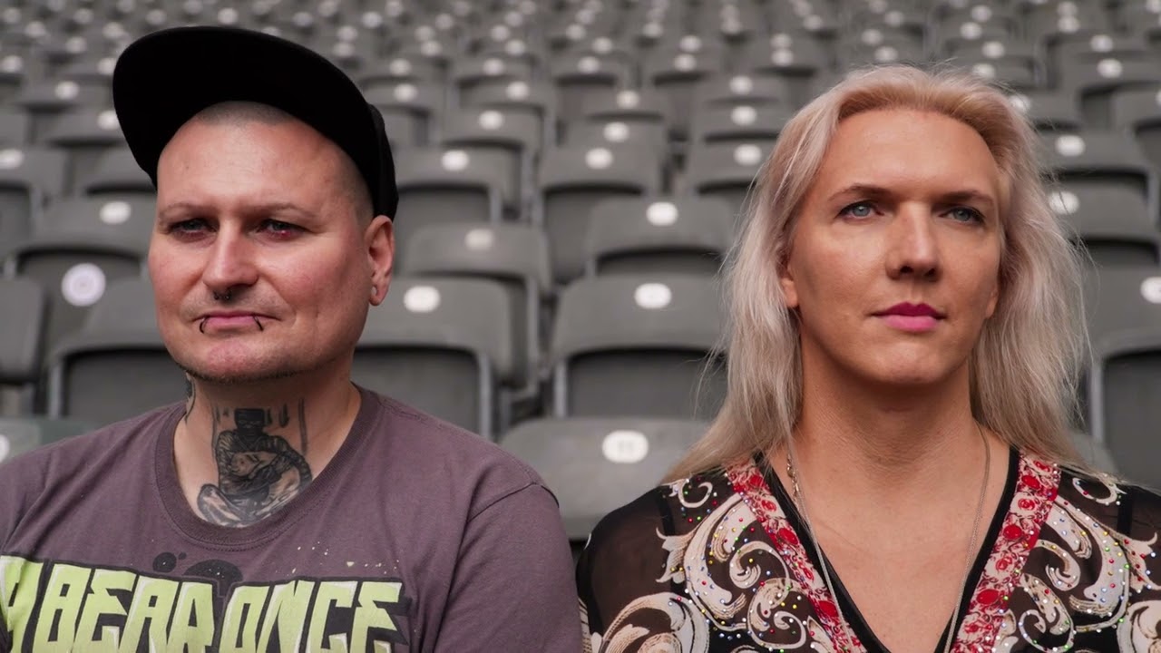 Auf dem Bild sind zwei Personen bis zu ihrer Brust abgebildet. Hinter ihnen sieht man leere Stuhlreihen, die mutmaßlich zu einem Fußballstadion gehören. Das Bild gehört zum Beitrag Vielfalt im Stadion - Queere Fans